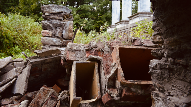 Collapsed chimney liners and brick, with freestanding white columns in the background, in a wooded setting.
