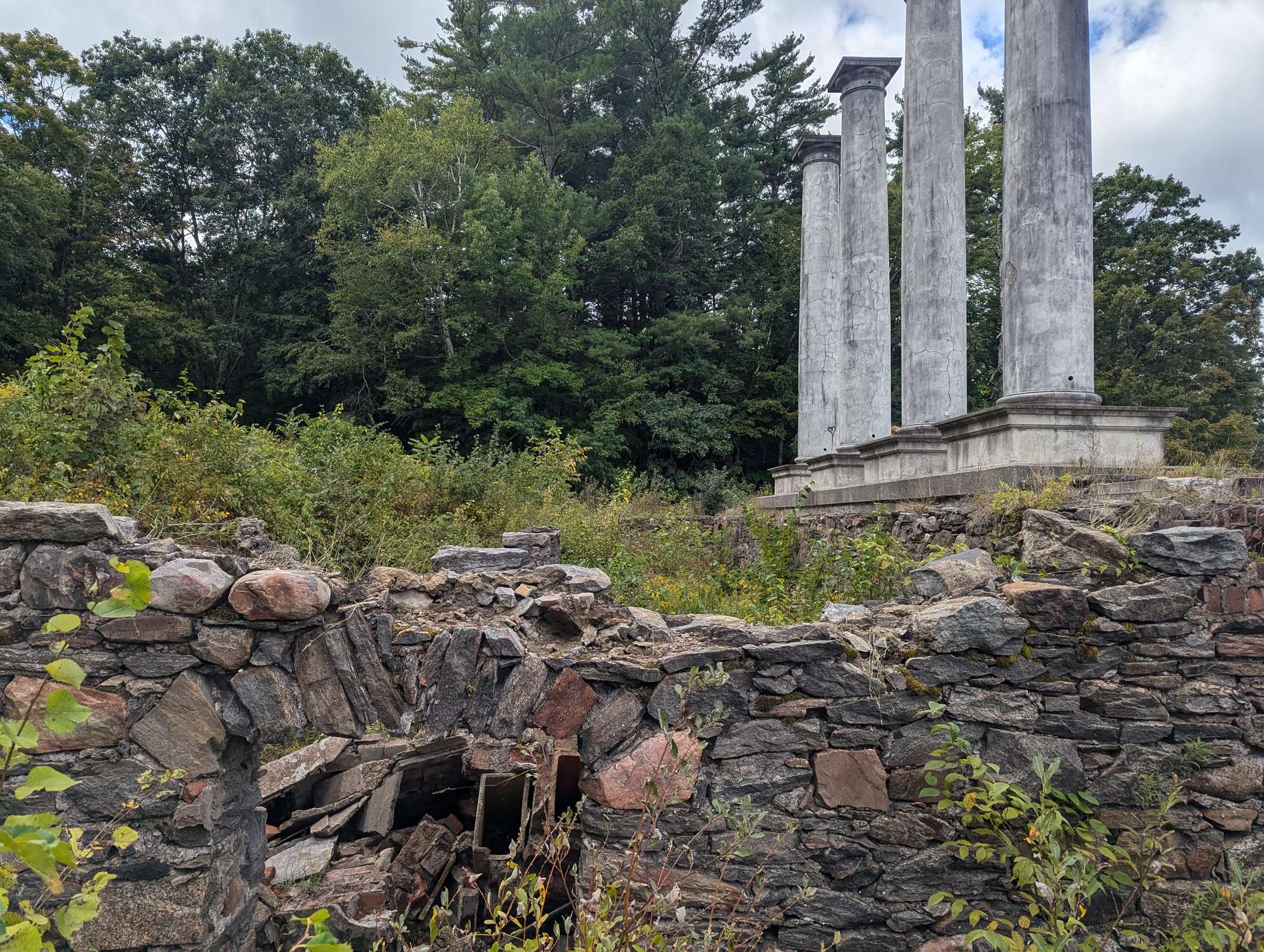 Four tall white columns sit atop a crumbling foundation, surrounded by forest.